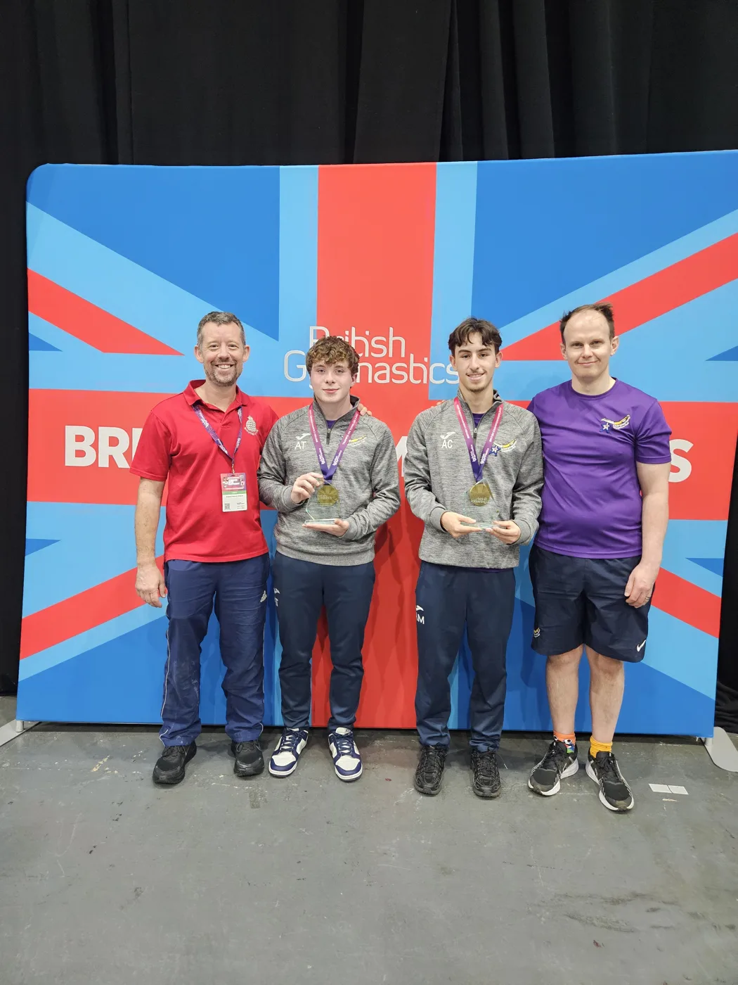Two young trampolinists in grey club tops holding gold medals and glass trophies, standing between two coaches in front of a Union Jack backdrop with the British Gymnastics logo